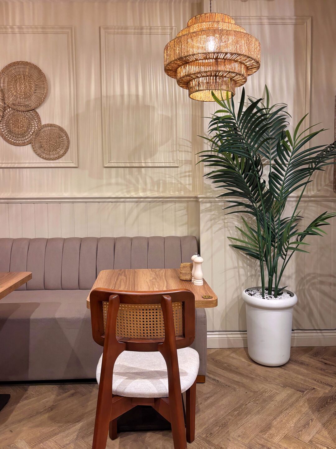 A banquette, a wooden table and a cane chair under a woven pendant light, with a tall palm in the corner